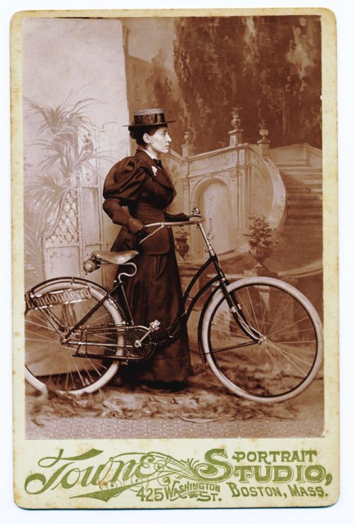 A woman in long Victorian dress, holding a bicycle.