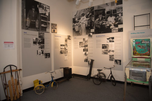 A museum wall with historic peices, including two tricycles, a pinball machine, and wall-mounted displays