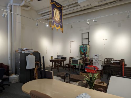 A room filled with a mess of file cabinets, tables, boxes, etc. The walls show marks where exhibits used to be