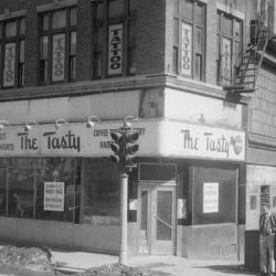 A store on the corner with a large script sign reading The Tasty, Hot Frankfurts, Coffee