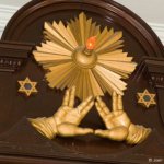 Two hands form a triangle under a radiant sun flanked by 2 stars of David on an ornate wood carving