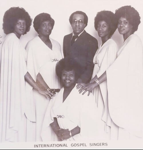 Five women in white dresses and a man in a suit and wearing glasses