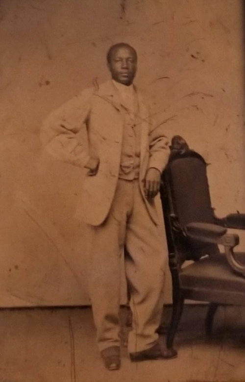 A black man standing firmly and looking directly into the camera wearing a light colored suit