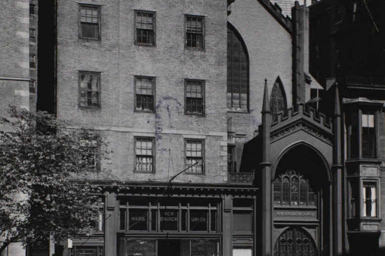Photograph of a five story brick building with a glass store front on the first floor. The building abuts a street.
