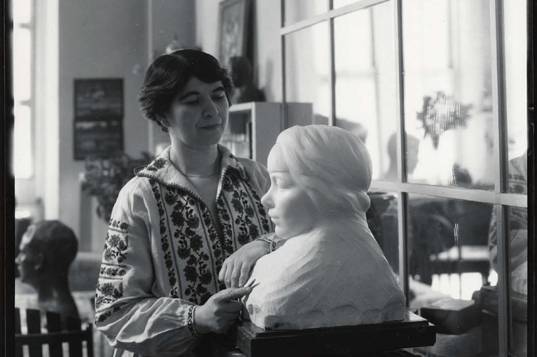 Bashka Paeff in her Studio Photograph of a woman in a dress with short dark hair standing in a window lit room next to a sculpture of a bust of a woman.