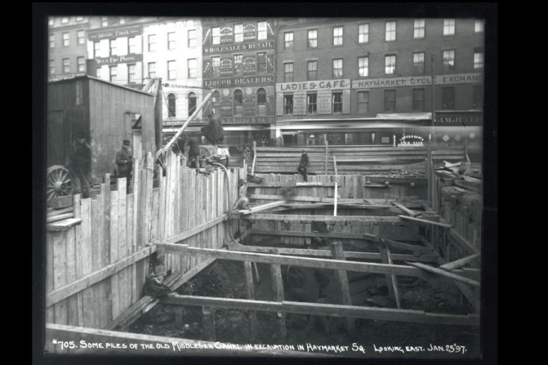 Photograph of a row of multi-story brick buildings with a dug out area of earth framed by wooden boards with workmen around it.