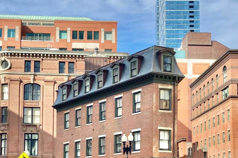 Photograph of the front of a four story brick building at an intersection with cars parked on the right side. Other