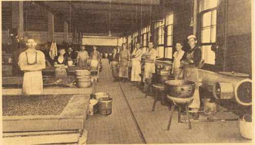 Photograph of twelve male workers standing in a semi-circle on a factory floor surrounded by equipment