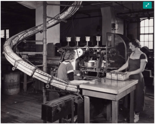 Photograph of two women, one sitting in front of machinery, the other standing near a counter, working to pack biscuit boxes. To the left of the women is a curved slide on which boxes moved toward the women.