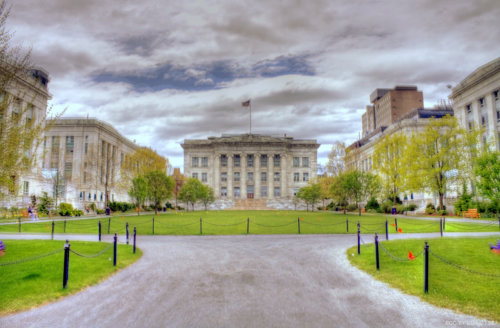 Photograph of a "U" shaped complex of similarly designed three-story stone buildings, the center of which is fronted by six columns on either side of its main entrance. The buildings surround a grass common and dirt path.