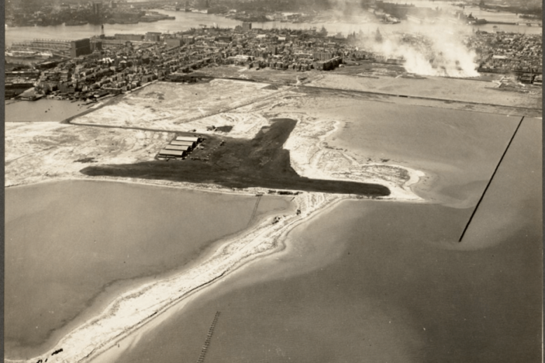 Aerial photograph of an airfield with four hangars and parked aircraft and vehicles, surrounded by water and and isthmus at the bottom and cleared land followed by a built up urban area at the top