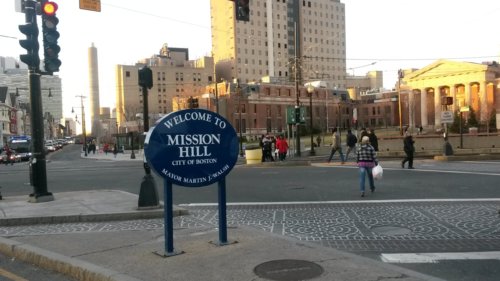 Photograph of a street intersection with residential housing and hospital buildings in the background and a sign stating, "Welcome to Mission Hill" in the foreground.