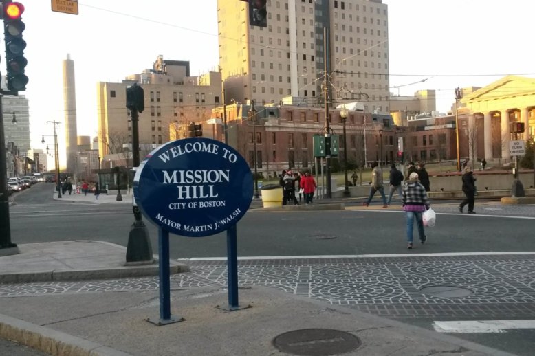 Photograph of a street intersection with residential housing and hospital buildings in the background and a sign stating, "Welcome to Mission Hill" in the foreground.