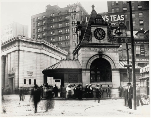 Photograph of a masonry subway station with a pitched tile roof and a clock. It is surrounded by a variety of brick and masonry building and pedestrians, some standing in front of the station. "Scollay Sq. Station" is chiseled across the front of the station building.