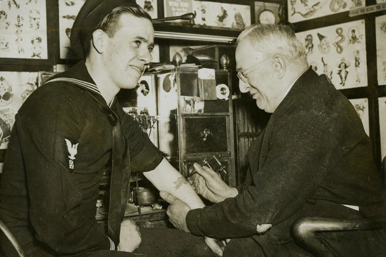 Photograph of a man on left in naval uniform having his left arm tattooed by an older man in glasses. Images of tattoo samples line the walls around them.