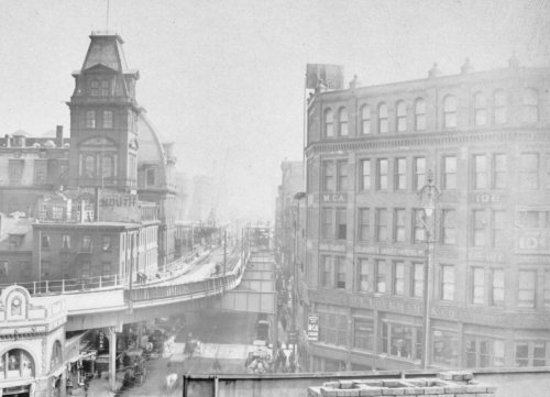 Photograph of a city square with brick and masonry buildings of varying sizes surrounding an elevated railroad track which is under construction. The building on the right bears sing "W.L. Lockhart & Co."