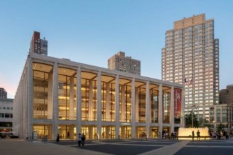 Photograph of a four-story glass and masonry building on a city square. The square is surrounded by apartment towers and people are walking about.