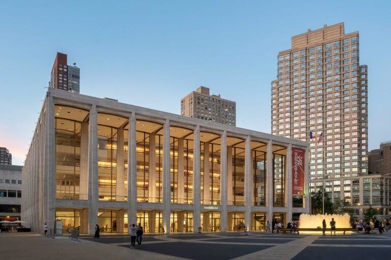 Photograph of a four-story glass and masonry building on a city square. The square is surrounded by apartment towers and people are walking about.
