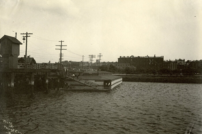 Photograph of a one-story structure sitting in the water beside a bridge.