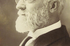 Photograph of a man with white hair, beard and eyeglasses wearing a suit and tie and sitting in profile.