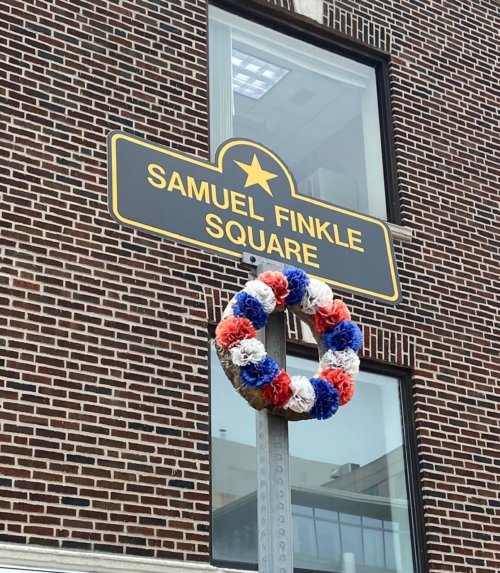 Photograph of a pole topped by a sign with a gold star and the words Samuel Finkle Square. A wreath of flowers is placed below the sign.
