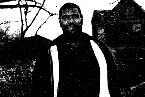 John_Moore Image from a newspaper articles showing a man with short hair and a beard wearing a jacket. He is standing in a wooded area with a two-story wooden house in the background.