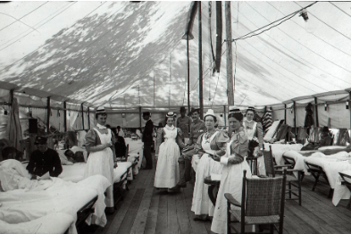 MGH Tent Ward Photograph of a tent lined with beds on the left and right, some occupied by patients. Five nurses in long dresses, with aprons, and a cap stand in between the beds, along with three soldiers.