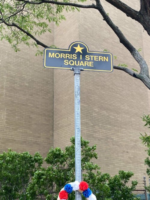 Photograph of a pole topped by a sign with a gold star and the words Morris I. Stern. A wreath of flowers is placed below the sign. A large multi-story brick building stands in the background.