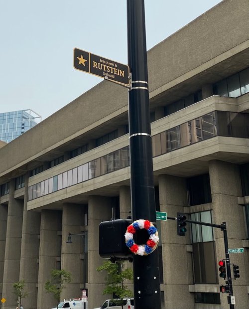Photograph of a pole topped by a sign with a gold star and the words Benjamin H Rutstein. A wreath of flowers is placed below the sign. A large multi-story concrete building stands in the background.