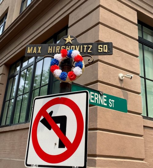 Photograph of a pole topped by a sign with a gold star and the words Max Hirshovitz . A wreath of flowers is placed below the sign.