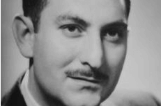 Photograph of a man with a mustache posing and wearing a suit and tie