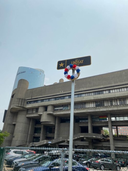 Photograph of a pole topped by a sign with a gold star and the words John F. Lindsay. A wreath of flowers is placed below the sign. A large multi-story concrete building stands in the background.