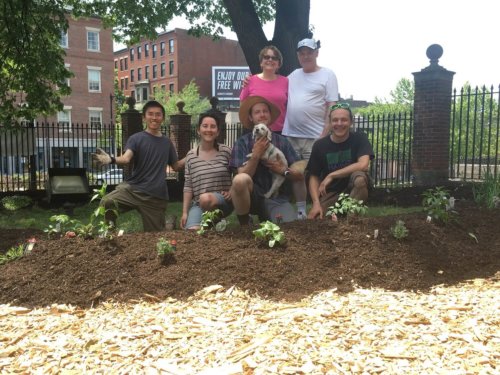 Photograph of six people posing in back of freshly planted flowers. Behind them is a fence, the street and brick buildings on the opposite side.