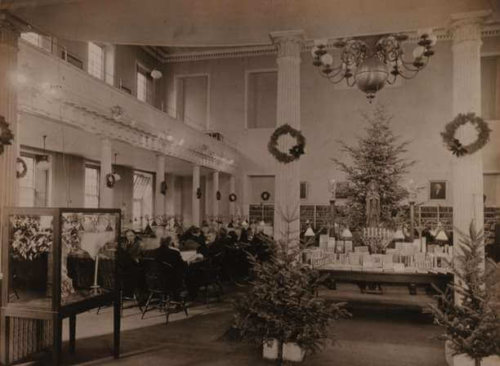 photograph of the interior of a library. A table decorated with winter holiday decorations sits between two columns in the center of the room. A second floor gallery is on the right, below which sit number of people in chairs reading.