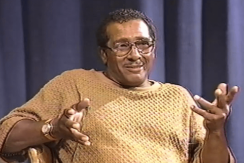 Photograph of a man wearing glasses and sweater sitting in a chair and gesturing with his hands.