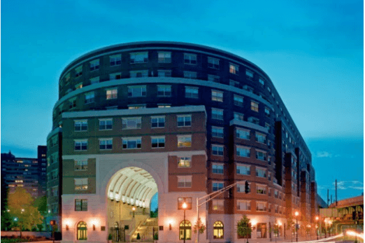 Photograph of the front of a U-shaped, 10-story, brick and masonry building with windows lining each floor. The building is at a city intersection and surrounded by lamp posts.