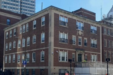 Photograph of the front of a three story brick and masonry building. A taller building stands to is right, and cars are parked in front on the street.