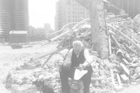 Photograph of an elderly man sitting with his dog next to a pile of stone and concrete debris.