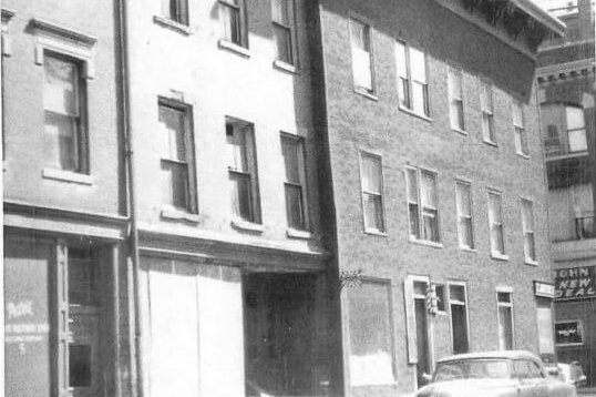 Photograph of a city block of three story brick buildings with cars parked along the curb