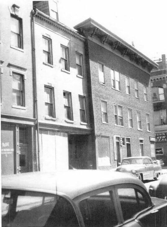 Photograph of a city block of three story brick buildings with cars parked along the curb