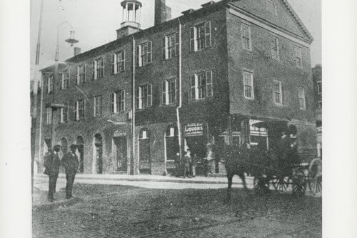 Photograph the front and side of a four-story brick building with a cupola. People horse drawn carts can be seen in the street in front.