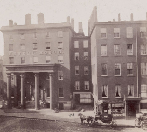 Photograph of a city block featuring the front of two joined brick, multi-story buildings lined with windows. The structure to the right has a front portico with four columns and the words "Revere House" attached above. Horse drawn buggies can be seen in the street.