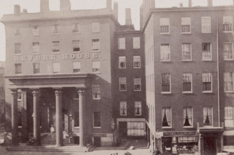 Photograph of a city block featuring the front of two joined brick, multi-story buildings lined with windows. The structure to the right has a front portico with four columns and the words "Revere House" attached above. Horse drawn buggies can be seen in the street.