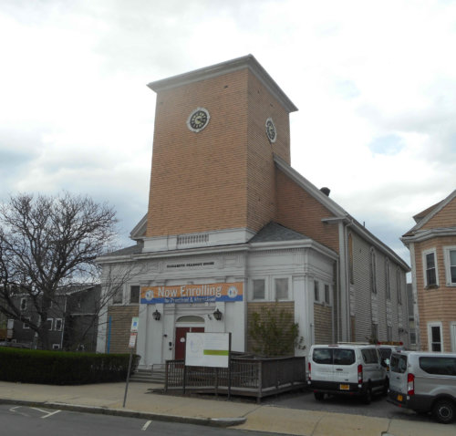 Photograph of the front of a wooden church with a clock but no steeple.