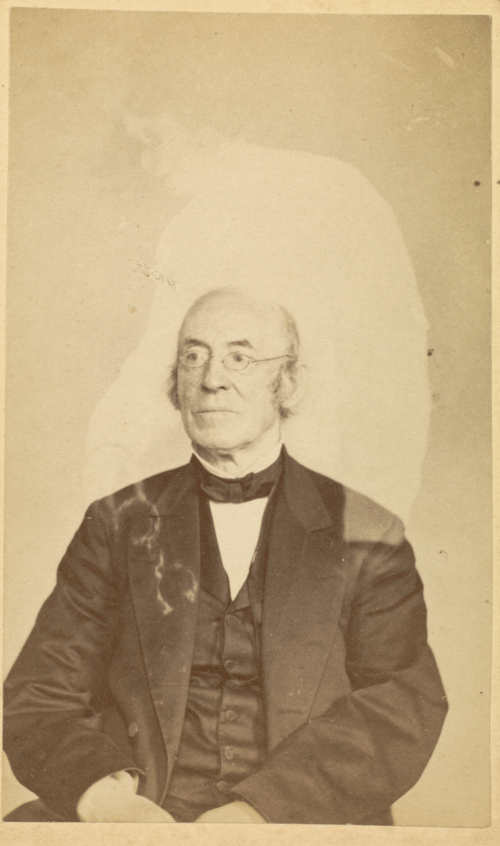 A man sits for a portrait with the faint outline of a person behind him.