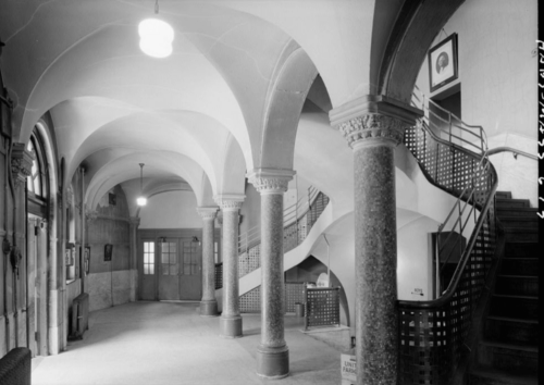 Black and white photograph of the inside of a school, showing an entryway with columns and a grand staircase.
