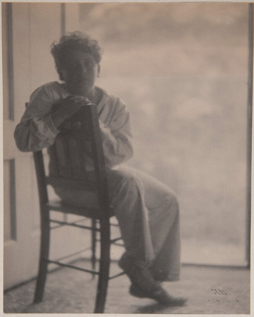 A photograph of an adolescent boy visible from head to toes sitting sideways on a chair in front of an open door.