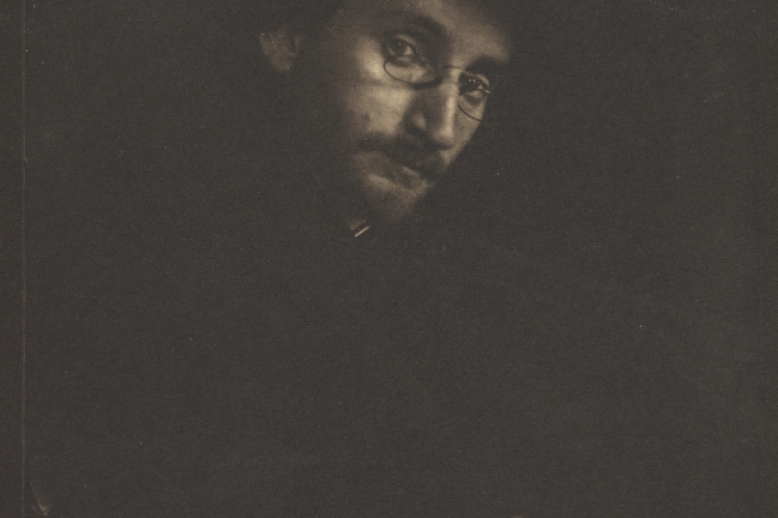 A photograph of a man in three-quarter profile. Nearly everything is in darkness except his face, turned to look at the camera, and his hands in the bottom right corner.