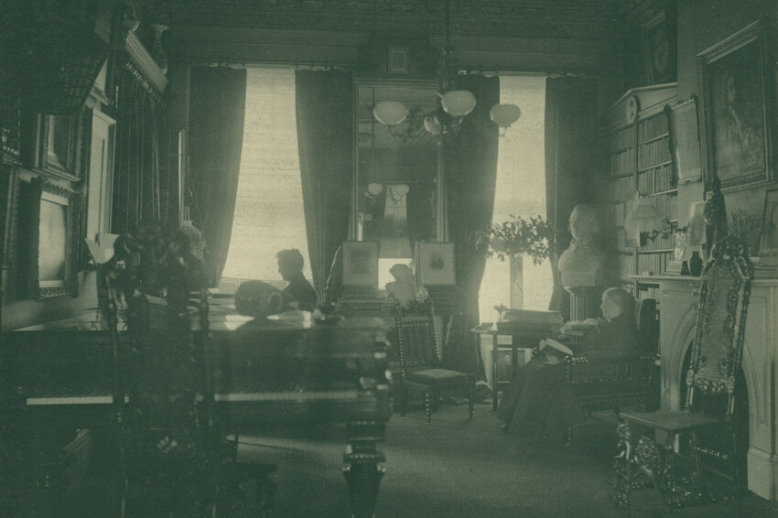 Sarah and Annie In a black and white photo, two women blend into a busy living room filled with a piano, chairs, sculptures, and art hanging on the walls.