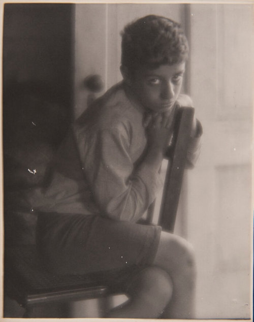 A photograph of an adolescent boy visible from the knees upward sitting backwards on a wooden chair, looking up at the viewer through his eyelashes.